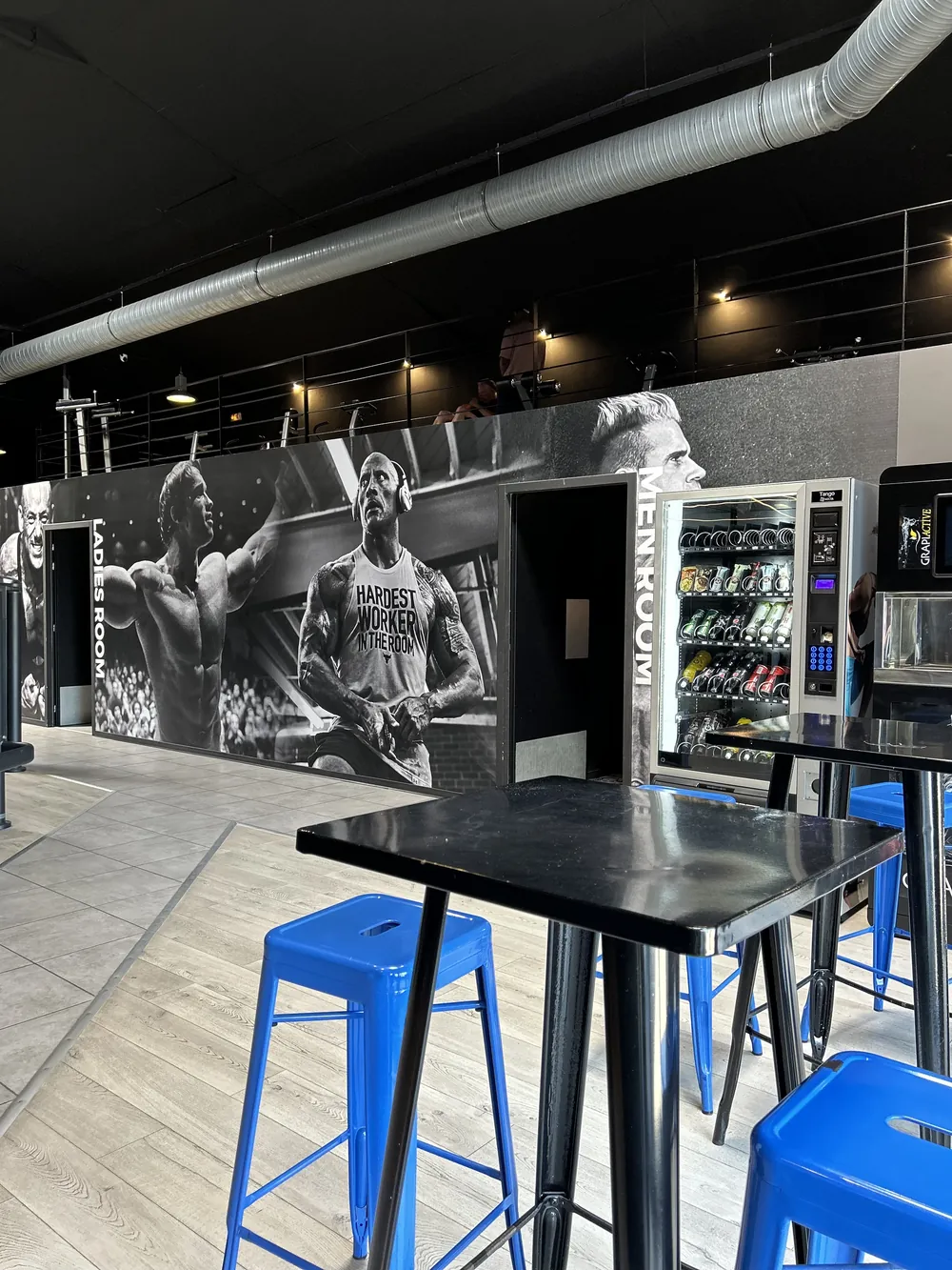 Coin détente du club avec tables hautes, tabourets bleus et fresque noir et blanc